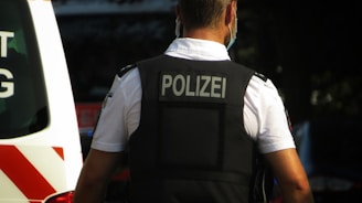 A police officer wearing a black vest with the word 'Polizei' on the back stands outdoors. The officer is facing away and has short hair, wearing a white shirt under the vest. Part of a vehicle with visible red and white stripes is on the left side of the image.