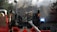 Firefighters in full gear work to control a house fire. A large fire truck with a ladder is positioned in front of a house, its roof engulfed in smoke and partially destroyed. The firefighters are using hoses to douse the flames, while smoke billows around them, and the emergency lights on the truck flash brightly.