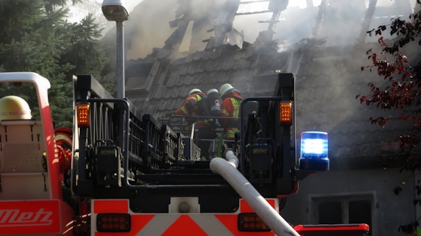 Firefighters in full gear work to control a house fire. A large fire truck with a ladder is positioned in front of a house, its roof engulfed in smoke and partially destroyed. The firefighters are using hoses to douse the flames, while smoke billows around them, and the emergency lights on the truck flash brightly.