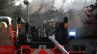 Firefighters in full gear work to control a house fire. A large fire truck with a ladder is positioned in front of a house, its roof engulfed in smoke and partially destroyed. The firefighters are using hoses to douse the flames, while smoke billows around them, and the emergency lights on the truck flash brightly.
