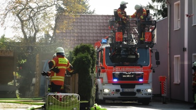 A firetruck is parked on a residential street with two firefighters standing on an elevated platform attached to the truck. Another firefighter is handling a hose in the background, near a wooden gate. The scene is set during the day with sunlight filtering through trees.