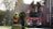 A firetruck is parked on a residential street with two firefighters standing on an elevated platform attached to the truck. Another firefighter is handling a hose in the background, near a wooden gate. The scene is set during the day with sunlight filtering through trees.