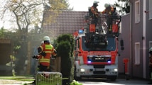 A firetruck is parked on a residential street with two firefighters standing on an elevated platform attached to the truck. Another firefighter is handling a hose in the background, near a wooden gate. The scene is set during the day with sunlight filtering through trees.