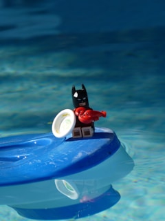 A small figurine of a character resembling a superhero with a black mask is standing on a blue floating object in water, holding a red lobster toy and a small white dish.