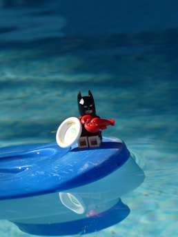 A small figurine of a character resembling a superhero with a black mask is standing on a blue floating object in water, holding a red lobster toy and a small white dish.