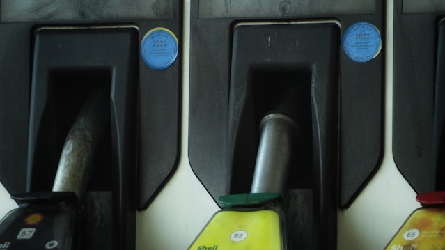 Three fuel pump nozzles are arranged in holders at a fueling station. The leftmost nozzle displays a Shell logo with 'B7', while the central nozzle is marked 'E5' with a green cap, and the right nozzle also shows 'E5' with a red cap. Blue stickers with the year 2022 are visible above each nozzle area, possibly indicating inspection stickers.