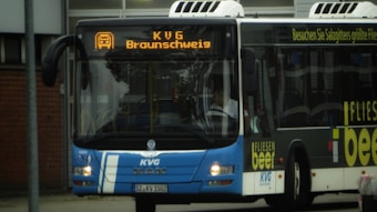 A city bus displaying the destination 'KVG Braunschweig' on its digital front sign. The bus is mainly blue with white accents, and there is an advertisement on its side. It is parked in an urban setting, and a person is visible inside, possibly the driver.