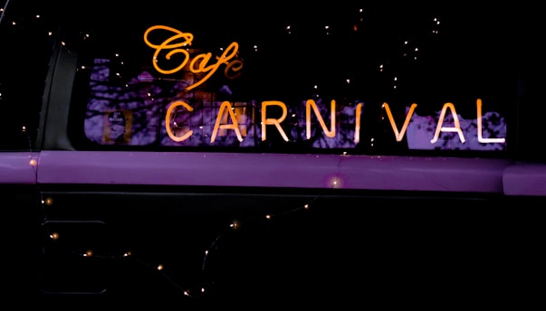 Warm neon lights spell out 'Cafe Carnival' against a dark background, accented by small twinkling fairy lights. The reflection of bare tree branches is faintly visible in the window.