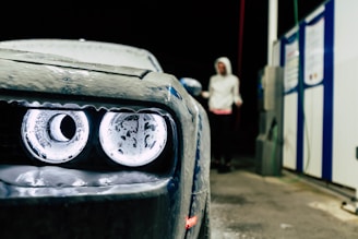 A close-up of a car's headlight covered in suds, with a person wearing a hooded sweatshirt standing in the background near a petrol station. The setting appears to be a car wash at night.