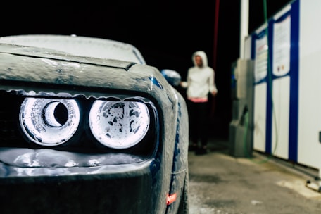 A close-up of a car's headlight covered in suds, with a person wearing a hooded sweatshirt standing in the background near a petrol station. The setting appears to be a car wash at night.
