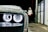 A close-up of a car's headlight covered in suds, with a person wearing a hooded sweatshirt standing in the background near a petrol station. The setting appears to be a car wash at night.