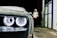A close-up of a car's headlight covered in suds, with a person wearing a hooded sweatshirt standing in the background near a petrol station. The setting appears to be a car wash at night.