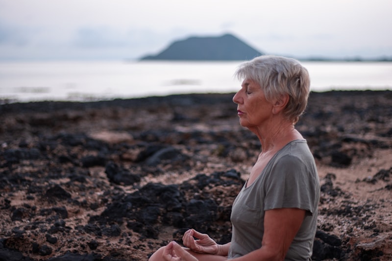 Woman in her 60s practicing seated meditation on a coastal rock at sunrise, calm and focused