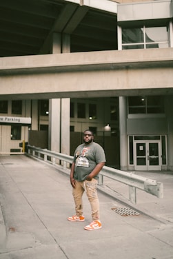 A person stands confidently in an urban environment. Wearing a graphic t-shirt, beige pants, and bright orange sneakers, the individual is positioned in a concrete area with industrial structures and parking signage in the background.