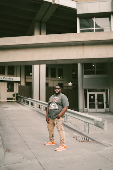 A person stands confidently in an urban environment. Wearing a graphic t-shirt, beige pants, and bright orange sneakers, the individual is positioned in a concrete area with industrial structures and parking signage in the background.