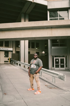 A person stands confidently in an urban environment. Wearing a graphic t-shirt, beige pants, and bright orange sneakers, the individual is positioned in a concrete area with industrial structures and parking signage in the background.