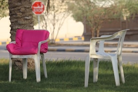A pair of white plastic chairs is positioned on a grassy lawn, with one featuring a bright pink cushion. The scene is set near a trunk of a palm tree, and a blurred stop sign is visible in the background along the street. The environment suggests a peaceful outdoor setting with soft lighting.