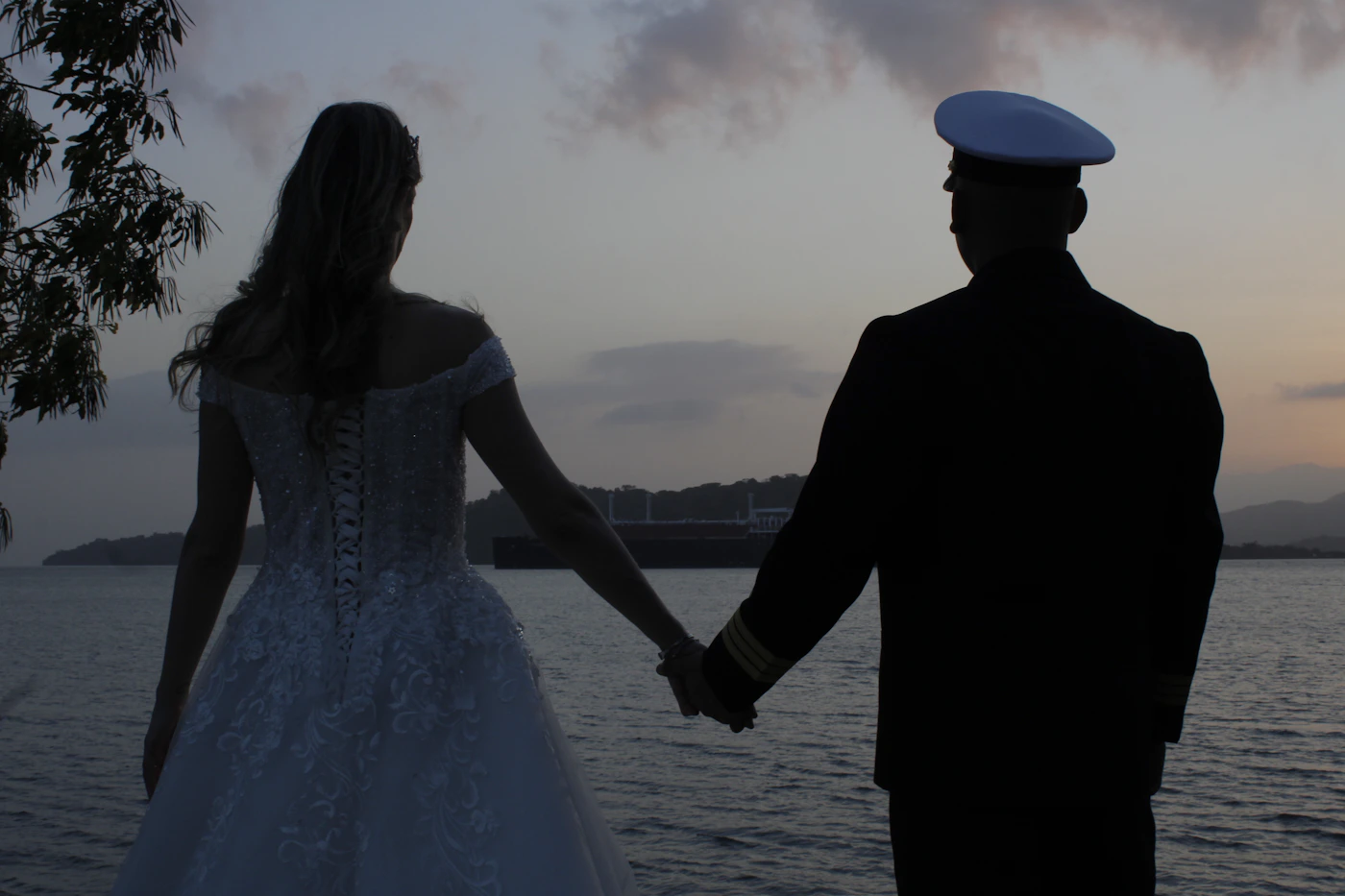 A couple holding hands walking along the path beside the Panama Canal with the historic canal locks and lush tropical vegetation visible behind them in the warm afternoon light