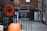 A blackboard sign with the opening hours 'Wed - Sat, 11 - 5' hangs on a metal fence. Beside it, a vibrant red wreath made of dried flowers is attached to the fence. In the foreground, there is an orange pumpkin placed on the ground. The background consists of a brick building and a paved alleyway, suggesting an urban setting.
