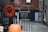 A blackboard sign with the opening hours 'Wed - Sat, 11 - 5' hangs on a metal fence. Beside it, a vibrant red wreath made of dried flowers is attached to the fence. In the foreground, there is an orange pumpkin placed on the ground. The background consists of a brick building and a paved alleyway, suggesting an urban setting.