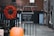 A blackboard sign with the opening hours 'Wed - Sat, 11 - 5' hangs on a metal fence. Beside it, a vibrant red wreath made of dried flowers is attached to the fence. In the foreground, there is an orange pumpkin placed on the ground. The background consists of a brick building and a paved alleyway, suggesting an urban setting.