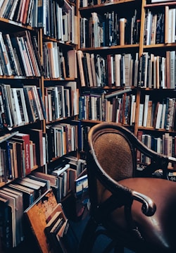 A cozy corner filled with bookshelves packed with a large variety of books. The books are arranged in an orderly fashion, and a vintage armchair sits invitingly in the foreground, its wooden frame and leather upholstery adding warmth to the scene.
