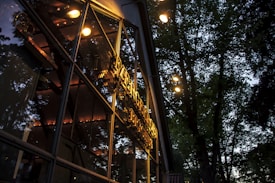 The image captures the exterior of a building with reflective glass windows, displaying warm interior lights. Large trees with dense foliage surround the structure, their outlines visible in the dim ambient light of what appears to be dusk or dawn.