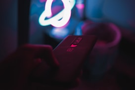 A dark room featuring a hand holding a smartphone with a prominent logo on the back. In the background, there are colorful neon lights forming abstract shapes.
