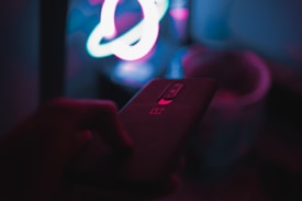 A dark room featuring a hand holding a smartphone with a prominent logo on the back. In the background, there are colorful neon lights forming abstract shapes.