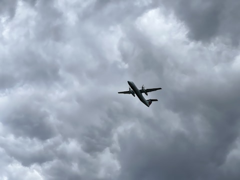 An airplane is flying upwards against a backdrop of thick, gray, and overcast clouds, indicating possible stormy weather.