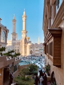 A bustling scene featuring a large mosque with towering minarets and intricate architecture surrounded by a crowd of people. The sky is clear and blue, providing a vibrant backdrop. Adjacent buildings display traditional design elements, and the space includes greenery and decorative elements such as fountains.