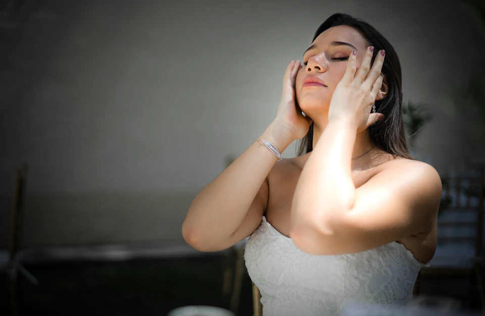 A bride in a white lace dress with her eyes closed, hands resting on her face, taking a quiet moment before the ceremony