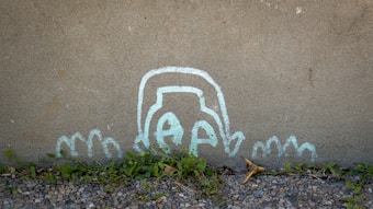 A whimsical chalk drawing of a simplified face partially submerged, with wavy lines that resemble hands or waves, on a concrete wall. The drawing is near the ground, with some green plants and small stones at the base of the wall.