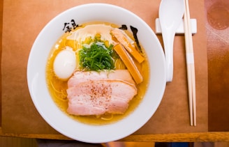 A bowl of ramen with rich, golden broth is served, containing sliced pork, a boiled egg, bamboo shoots, and green onions. It is accompanied by chopsticks and a spoon, placed on a wooden surface.