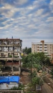A construction site with an unfinished building next to a residential area. The building shows exposed concrete and some bricks, with a couple of workers on the structure. In the foreground, a blue tarp covers some materials, and nearby, trees and electrical wires are visible. The sky is overcast with scattered clouds.