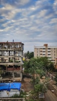 A construction site with an unfinished building next to a residential area. The building shows exposed concrete and some bricks, with a couple of workers on the structure. In the foreground, a blue tarp covers some materials, and nearby, trees and electrical wires are visible. The sky is overcast with scattered clouds.
