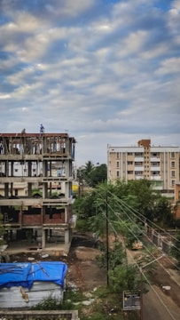 A construction site with an unfinished building next to a residential area. The building shows exposed concrete and some bricks, with a couple of workers on the structure. In the foreground, a blue tarp covers some materials, and nearby, trees and electrical wires are visible. The sky is overcast with scattered clouds.