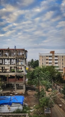 A construction site with an unfinished building next to a residential area. The building shows exposed concrete and some bricks, with a couple of workers on the structure. In the foreground, a blue tarp covers some materials, and nearby, trees and electrical wires are visible. The sky is overcast with scattered clouds.