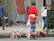 A person walks multiple small dogs on leashes, each wearing matching red jackets. There are other pedestrians in the background on a city street, with some store signs visible.