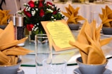 A round dining table set for an event with neatly folded beige napkins atop black bowls. The centerpiece includes a bouquet of red roses and white flowers in a clear vase. A yellow menu card rests prominently in the center. The table also features glassware and a salt and pepper shaker set.
