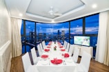 A luxurious dining room with a long table set for a formal dinner, featuring white tablecloths and red napkins folded elegantly. Large windows provide an expansive view of a city skyline and waterfront at dusk, with buildings illuminated against a deep blue sky. The room has white walls, ceiling lights, and a ceiling fan.
