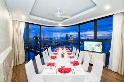 A luxurious dining room with a long table set for a formal dinner, featuring white tablecloths and red napkins folded elegantly. Large windows provide an expansive view of a city skyline and waterfront at dusk, with buildings illuminated against a deep blue sky. The room has white walls, ceiling lights, and a ceiling fan.