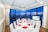 A luxurious dining room with a long table set for a formal dinner, featuring white tablecloths and red napkins folded elegantly. Large windows provide an expansive view of a city skyline and waterfront at dusk, with buildings illuminated against a deep blue sky. The room has white walls, ceiling lights, and a ceiling fan.