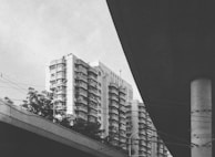 A high-rise residential building with multiple balconies and architectural details is surrounded by trees and framed by the dark structure of an overpass. Power lines are visible crossing the scene.