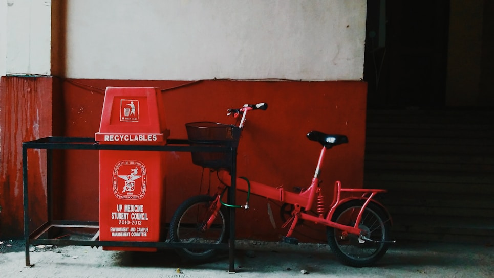 A red recycling bin labeled 'Recyclables' placed under a shelter with a red bicycle leaning against it. The bin has an inscription from the UP Medicine Student Council along with other text. The background shows a two-tone wall, with the lower part painted red and the upper part beige.