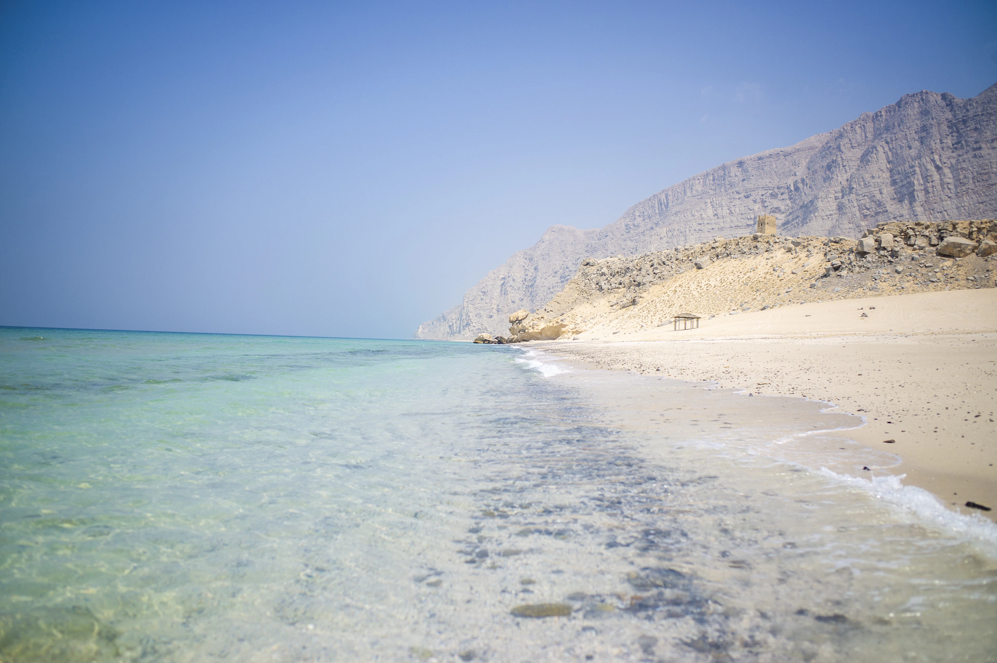 Clear turquoise water, a strip of sand and steep bare cliffs on the Musandam coast, Oman