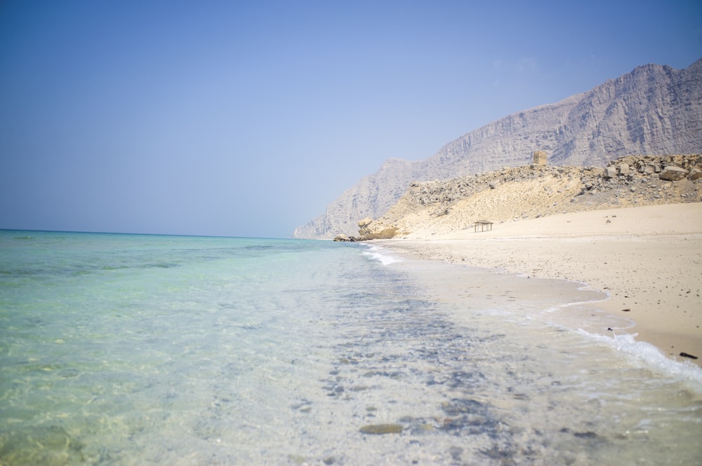 a sandy beach with clear blue water and mountains in the background