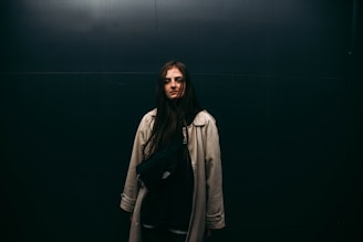 A person with long dark hair stands against a dark, minimalistic background wearing a light-colored trench coat and a black crossbody bag.