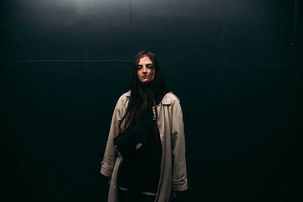 A person with long dark hair stands against a dark, minimalistic background wearing a light-colored trench coat and a black crossbody bag.