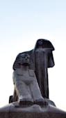 Large stone statues depicting an Egyptian sphinx and a robed woman standing behind it. The sphinx has a stoic expression with traditional Egyptian headgear, while the robed woman stretches one arm up, creating an impression of grace and authority. The background is a clear sky during daytime.